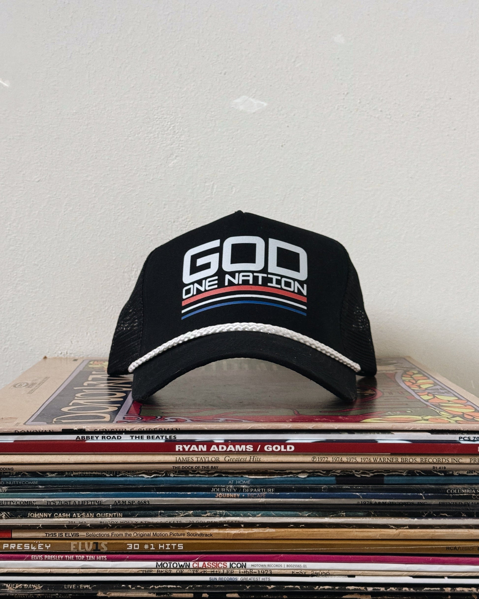 A black baseball cap with white text 'GOD ONE NATION' on the front, placed on a stack of vinyl records.
