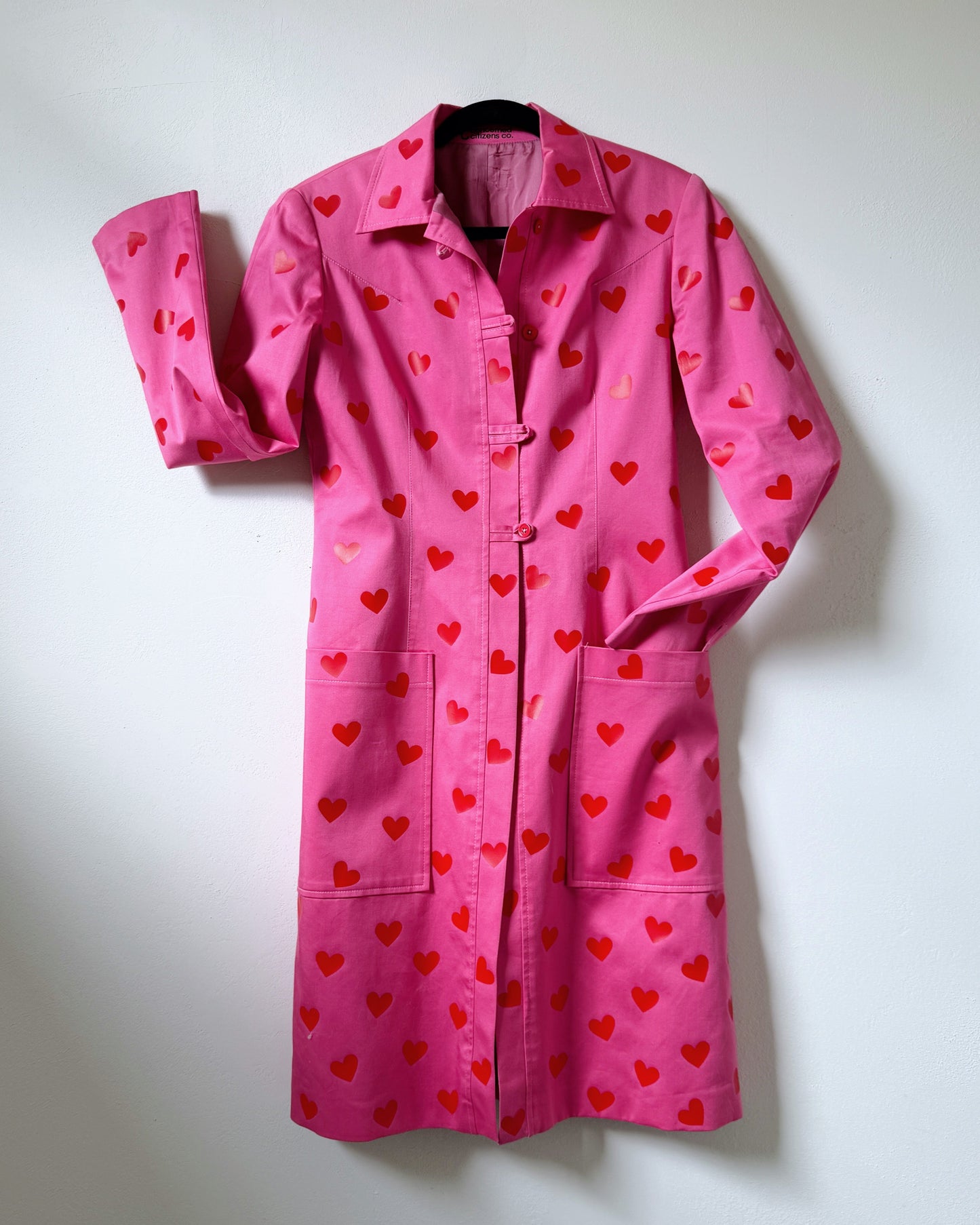 A pink trench coat with a pattern of red mini hearts displayed against a neutral background.