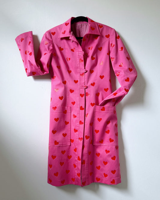 A pink trench coat with a pattern of red mini hearts displayed against a neutral background.