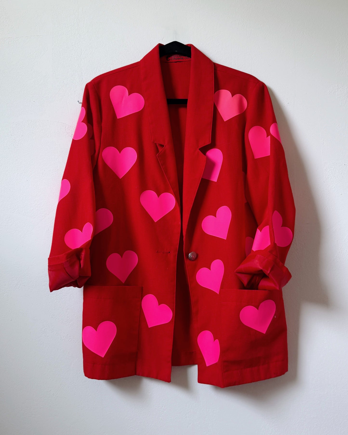 A red blazer with a pattern of neon pink hearts spread across the fabric, displayed against a white background. Upcycled womens jacket with bold neon pink hearts.