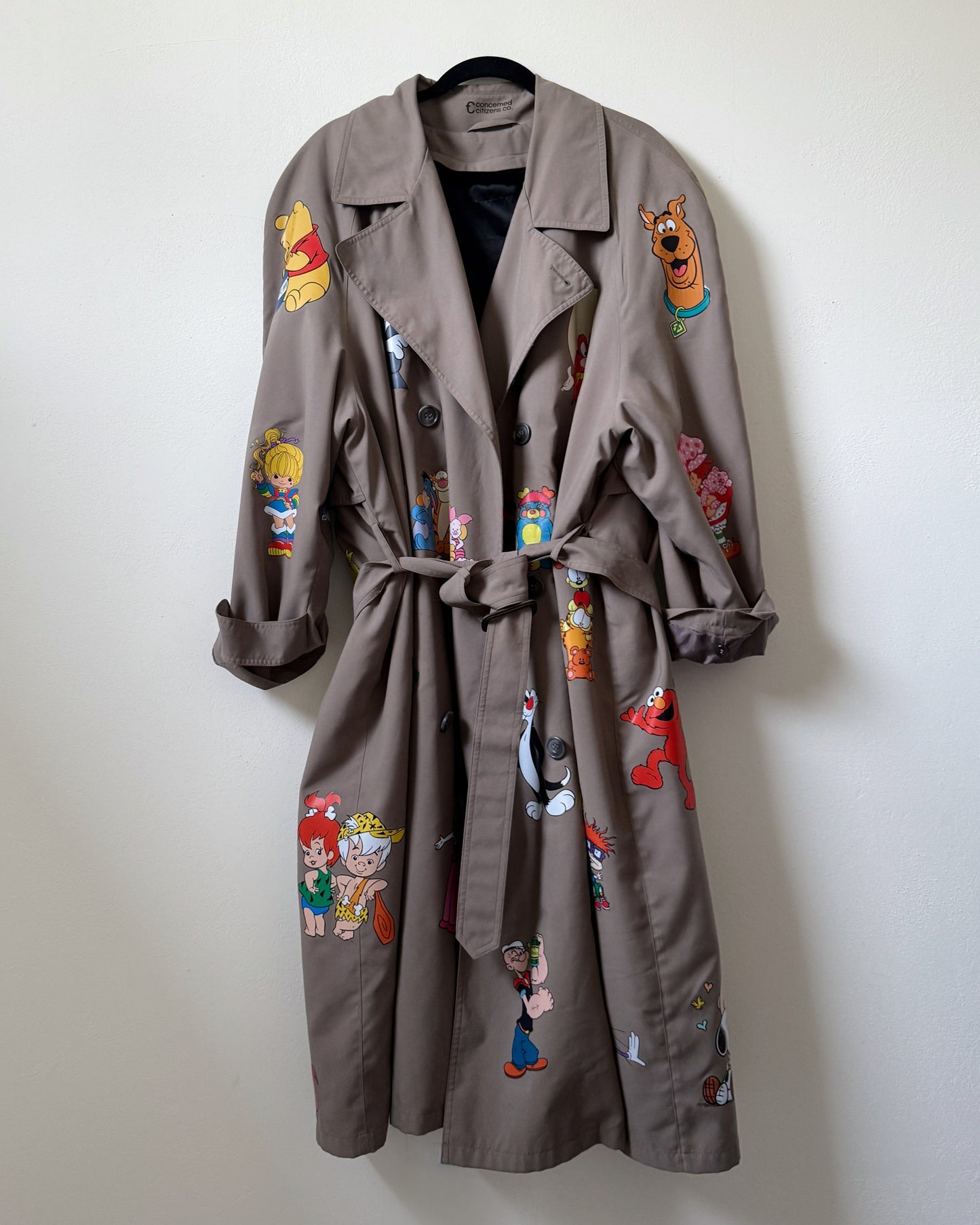 A pre-loved khaki/gray trench coat with vinyl cartoon character designs, displayed on a hanger against a plain wall.