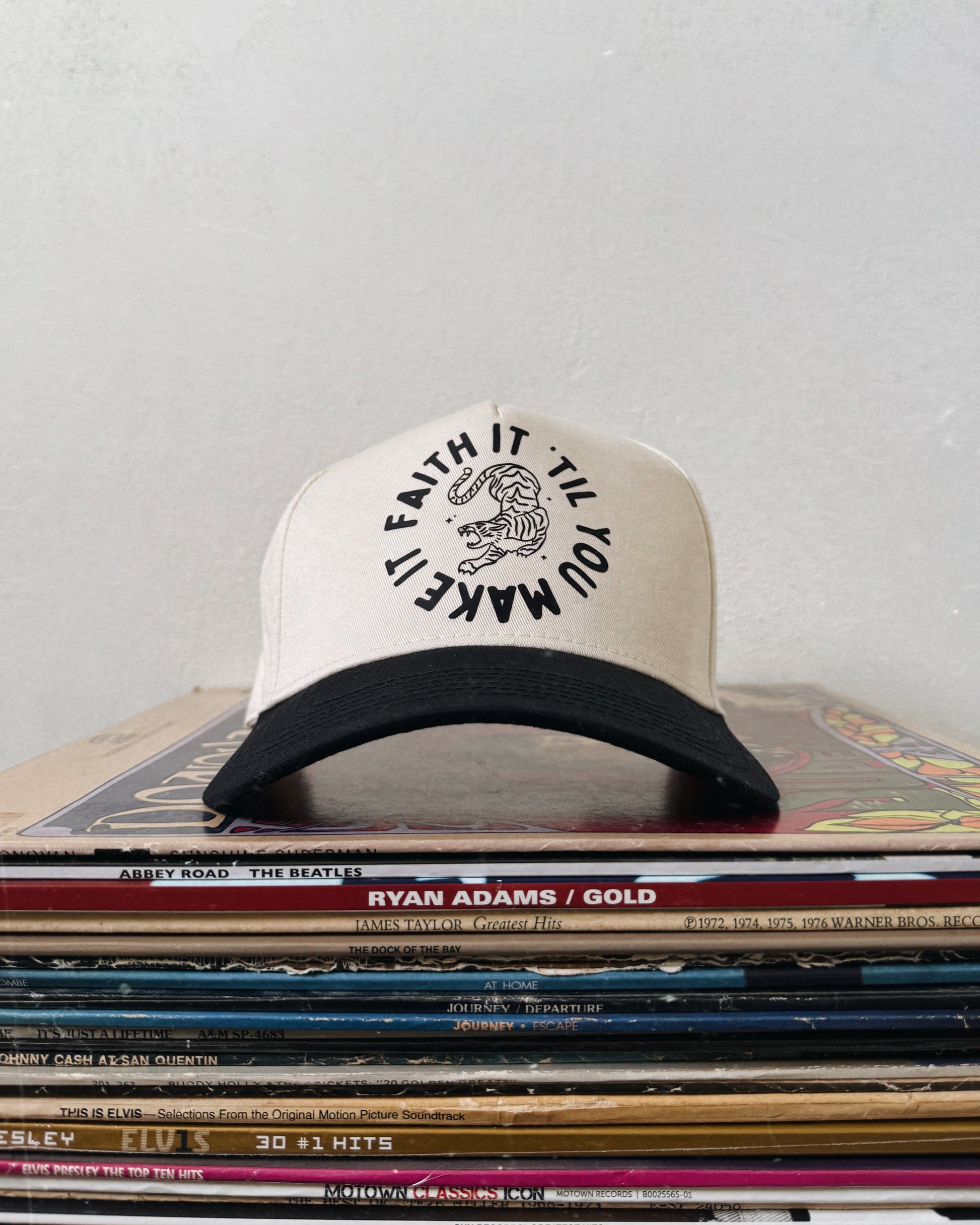A beige and black baseball hat with a text and logo on the front, placed on a stack of vinyl records.