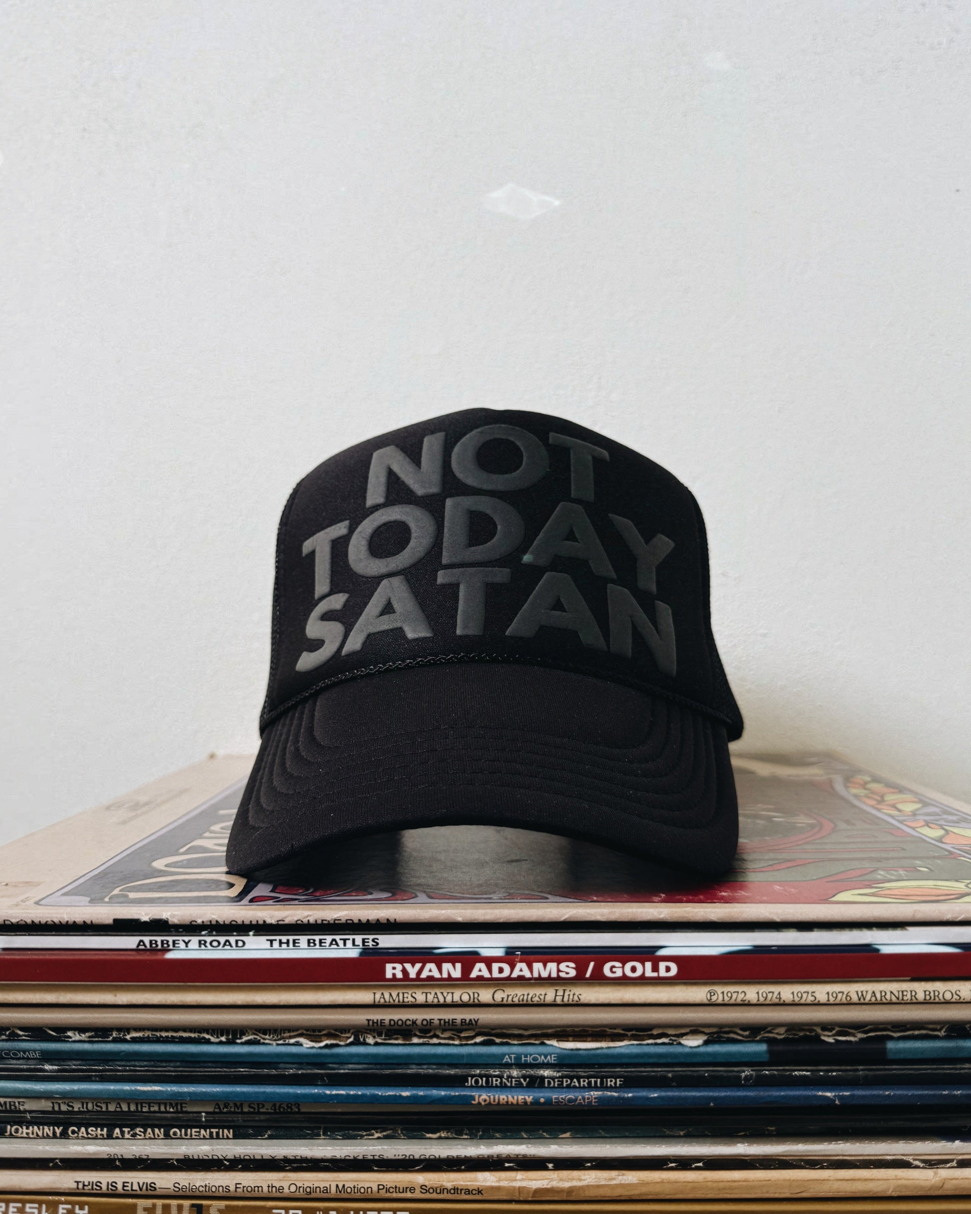 A black trucker hat with the text 'NOT TODAY SATAN' printed in white on the front, placed on a stack of vinyl records.