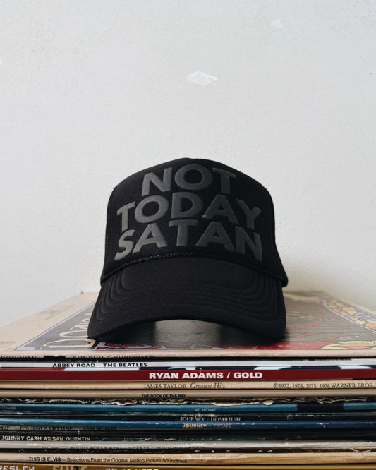A black trucker hat with the text 'NOT TODAY SATAN' printed in white on the front, placed on a stack of vinyl records.