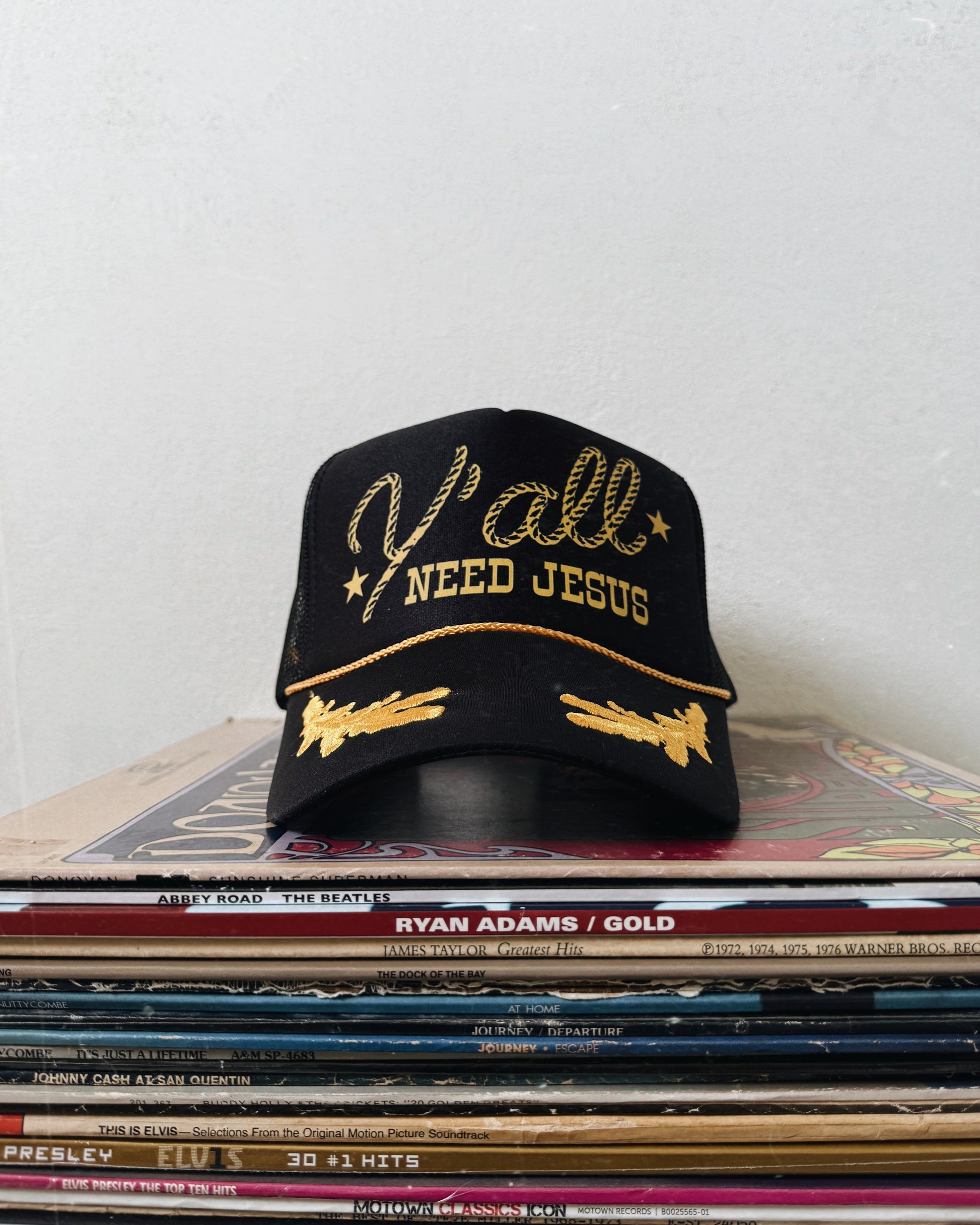 A black trucker hat with gold text and oak leaf patterns, placed on a stack of vinyl records, with the phrase 'Y'ALL NEED JESUS' embroidered on the front.