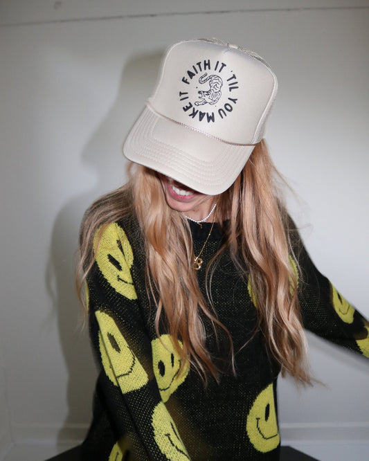 Person wearing a white cap with text and a black sweater with yellow smiley face patterns.