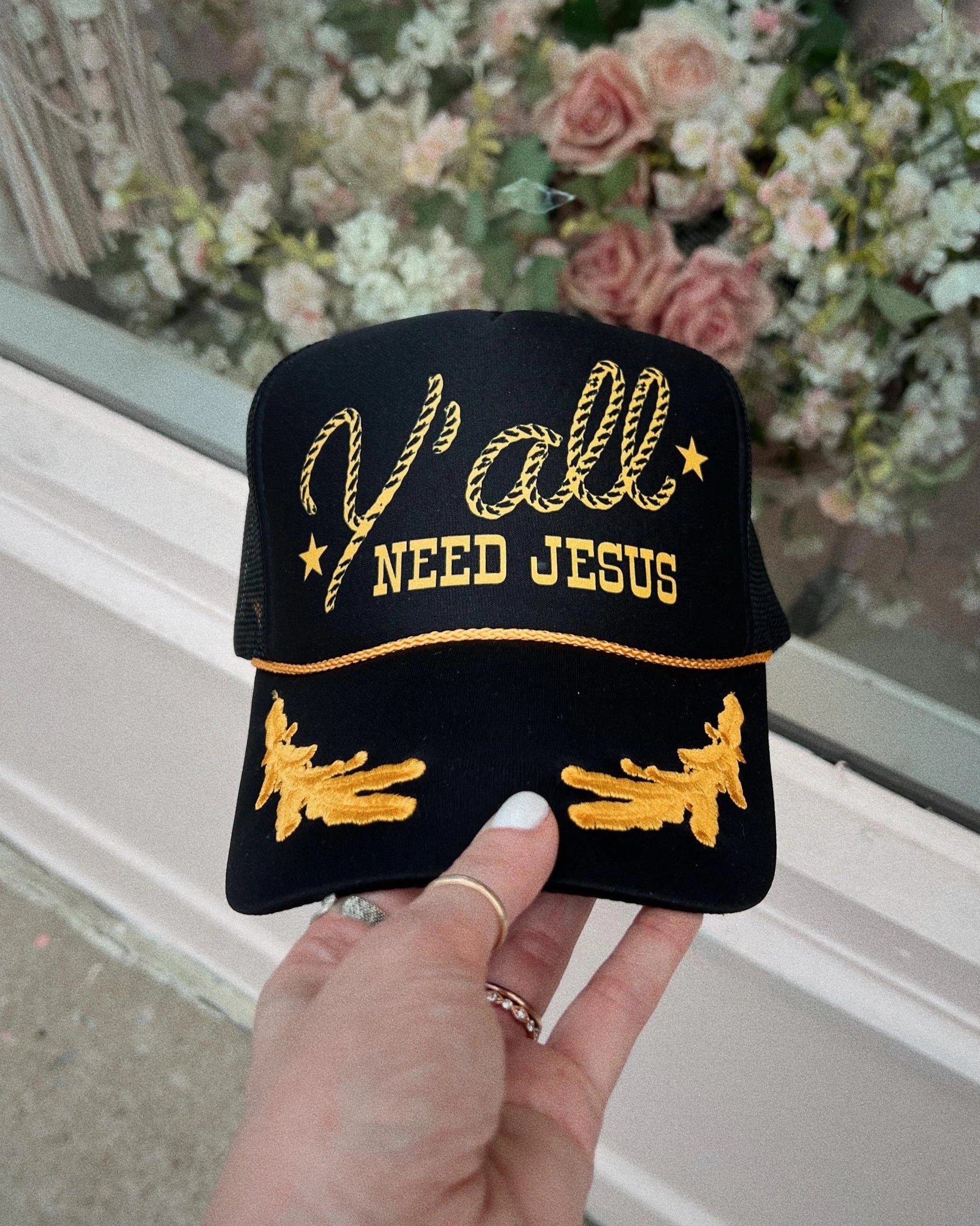 A black and gold trucker hat with the text 'Y'ALL NEED JESUS' and oak leaf designs held up against a backdrop of a floral arrangement.