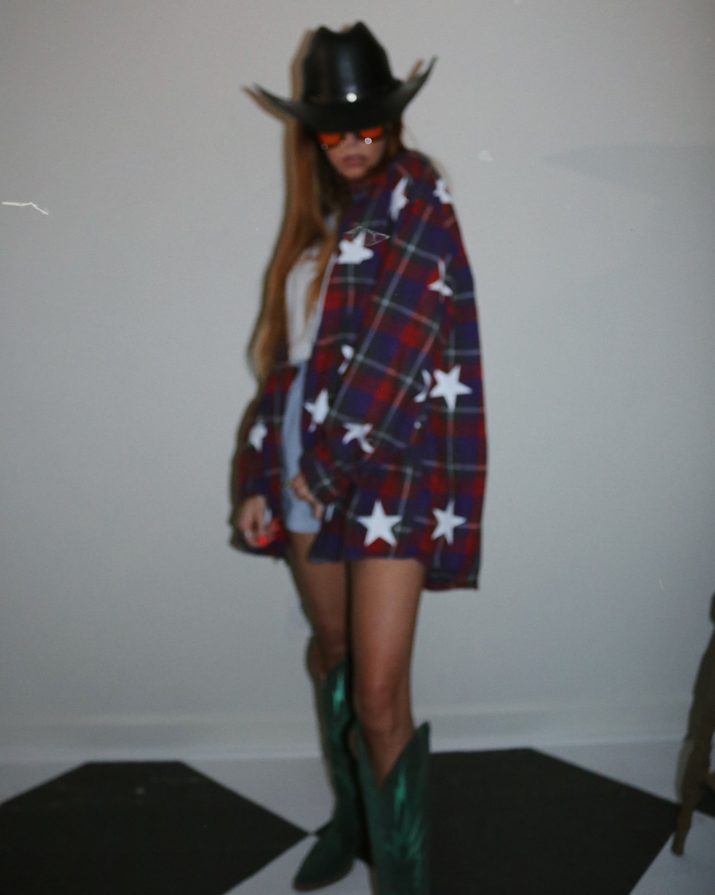Person wearing a plaid coat with star patterns, green boots, and a black cowboy hat indoors.