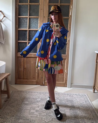 Person wearing a colorful outfit with a blue jacket and multicolored skirt in a room with wooden doors and a rug.