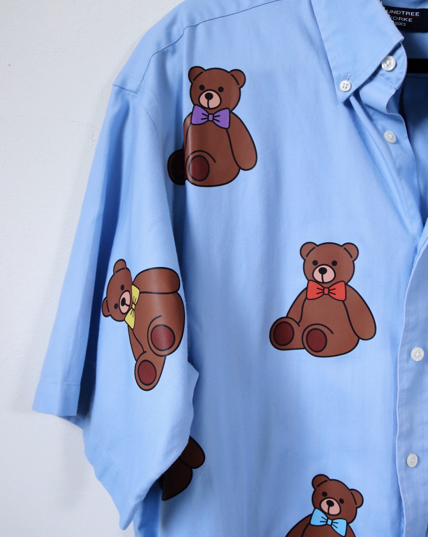 Blue shirt with brown teddy bear illustrations wearing colorful bow ties on a white background