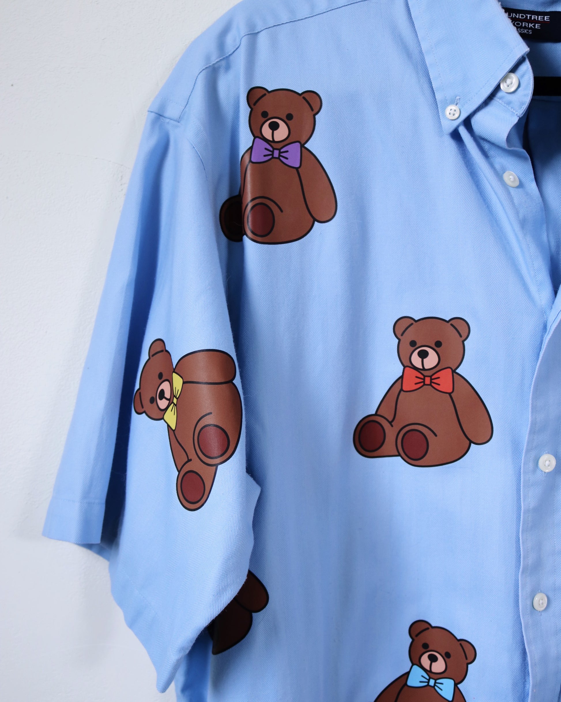 Blue shirt with brown teddy bear illustrations wearing colorful bow ties on a white background