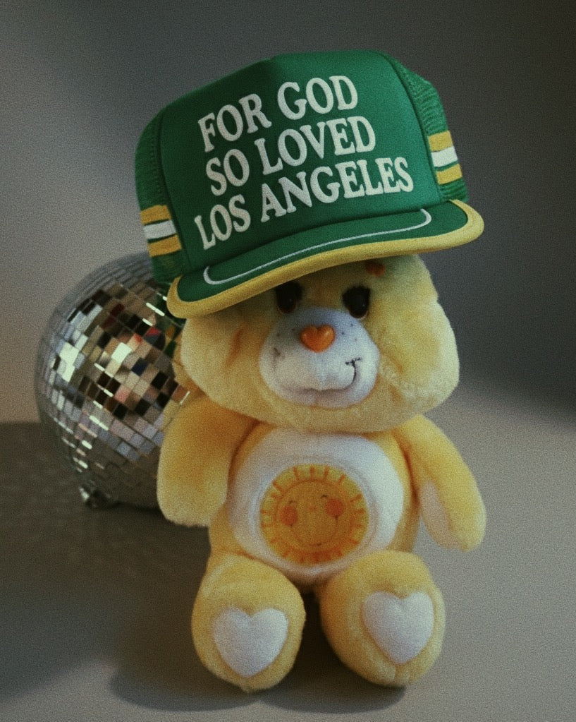 Plush bear wearing a green cap with text, sitting on a reflective surface.