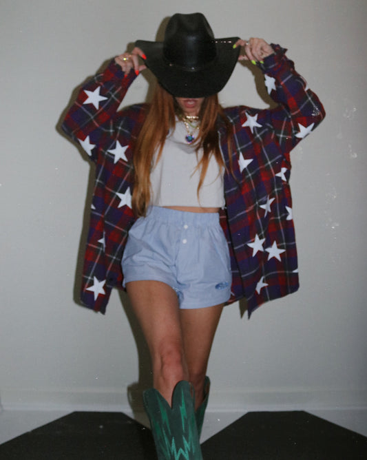 Person wearing a plaid shirt with star patterns, shorts, and cowboy boots against a plain background